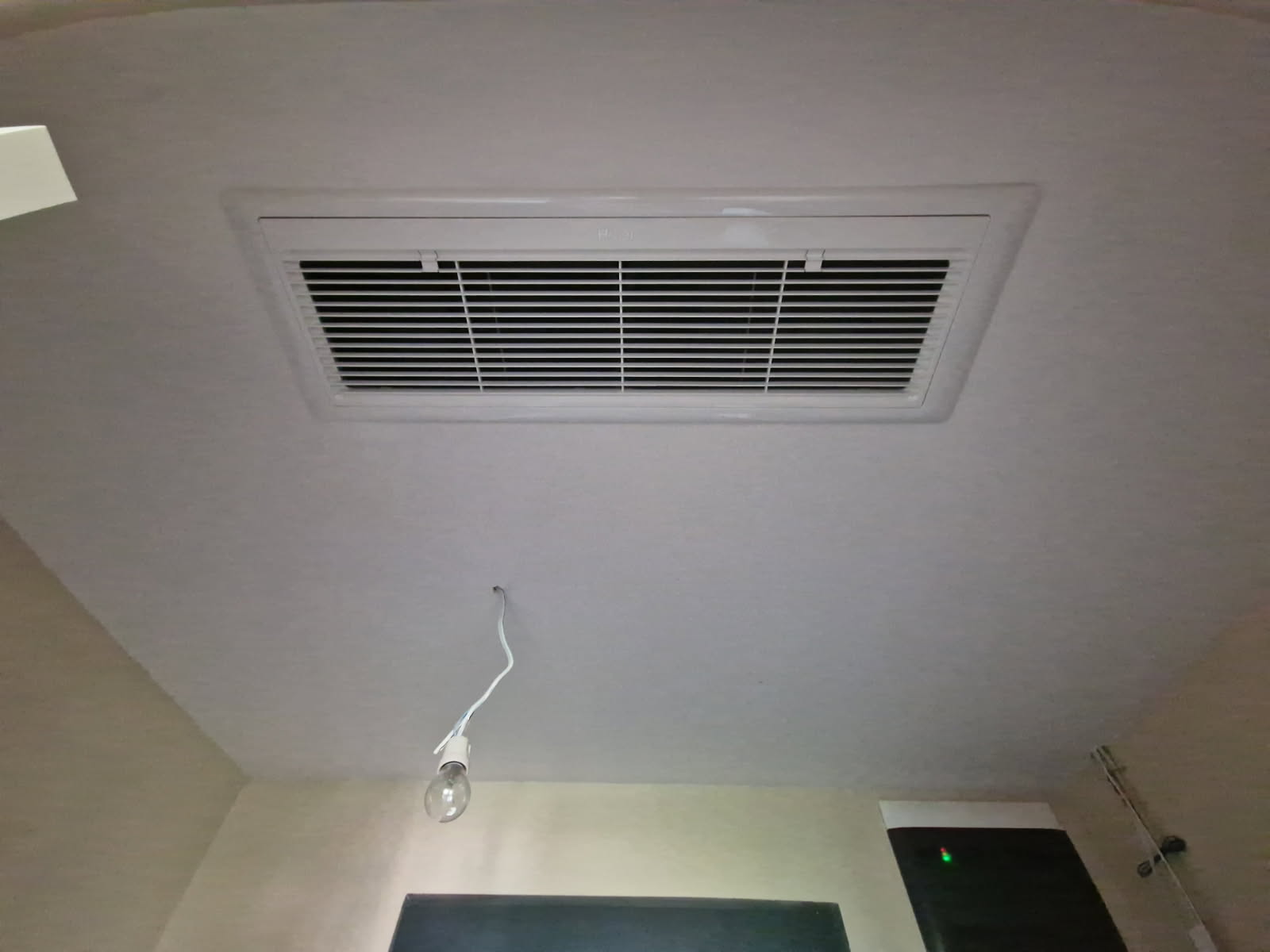 Zdjęcie przedstawia białą kratkę nawiewną (anemostat/nawiewnik) klimatyzacji kanałowej Haier Slim Duct, zamontowaną w podwieszanym suficie. Na pierwszym planie widoczny jest również wiszący przewód z żarówką.