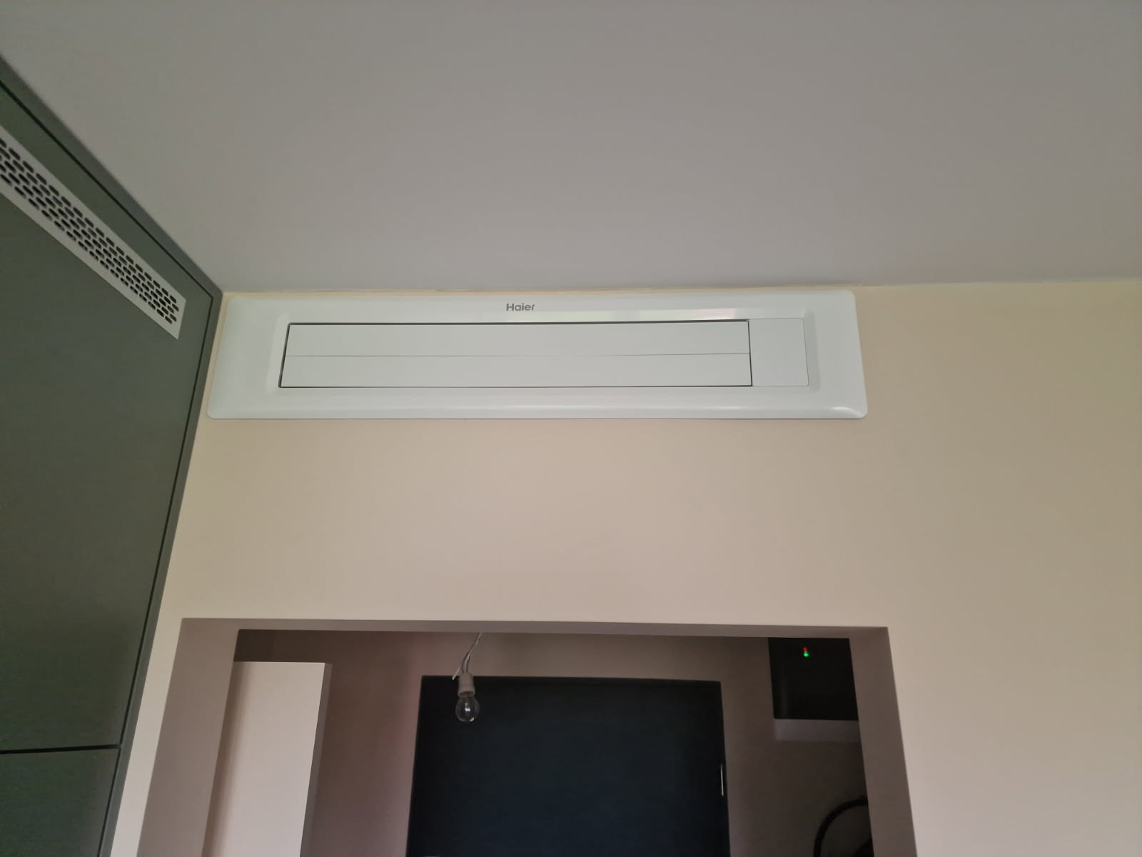 Zdjęcie przedstawia zamontowany panel ścienny/nawiewny z widocznym logo Haier, umieszczony w ścianie powyżej przejścia do innego pomieszczenia.